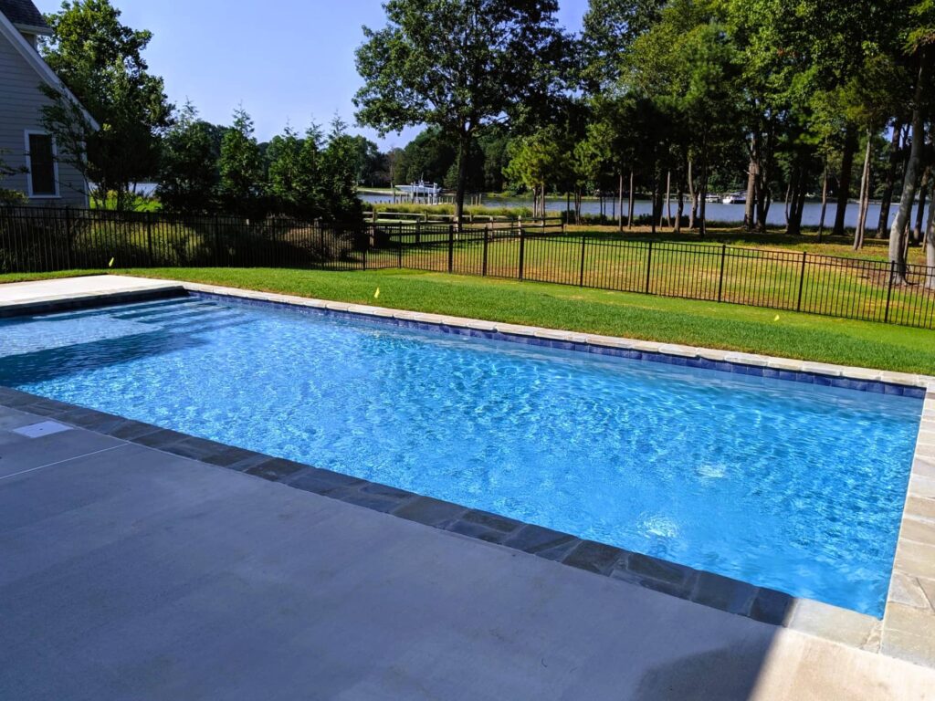 geometric pool featuring a tanning ledge next to grass area and fence